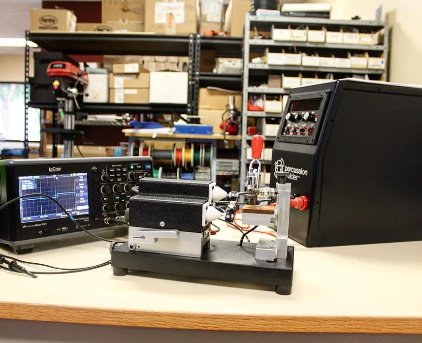 Percussion Welder 858 and 726 bench fixture beside a LeCroy oscilloscope, with wire spools and shelving behind on a lab bench