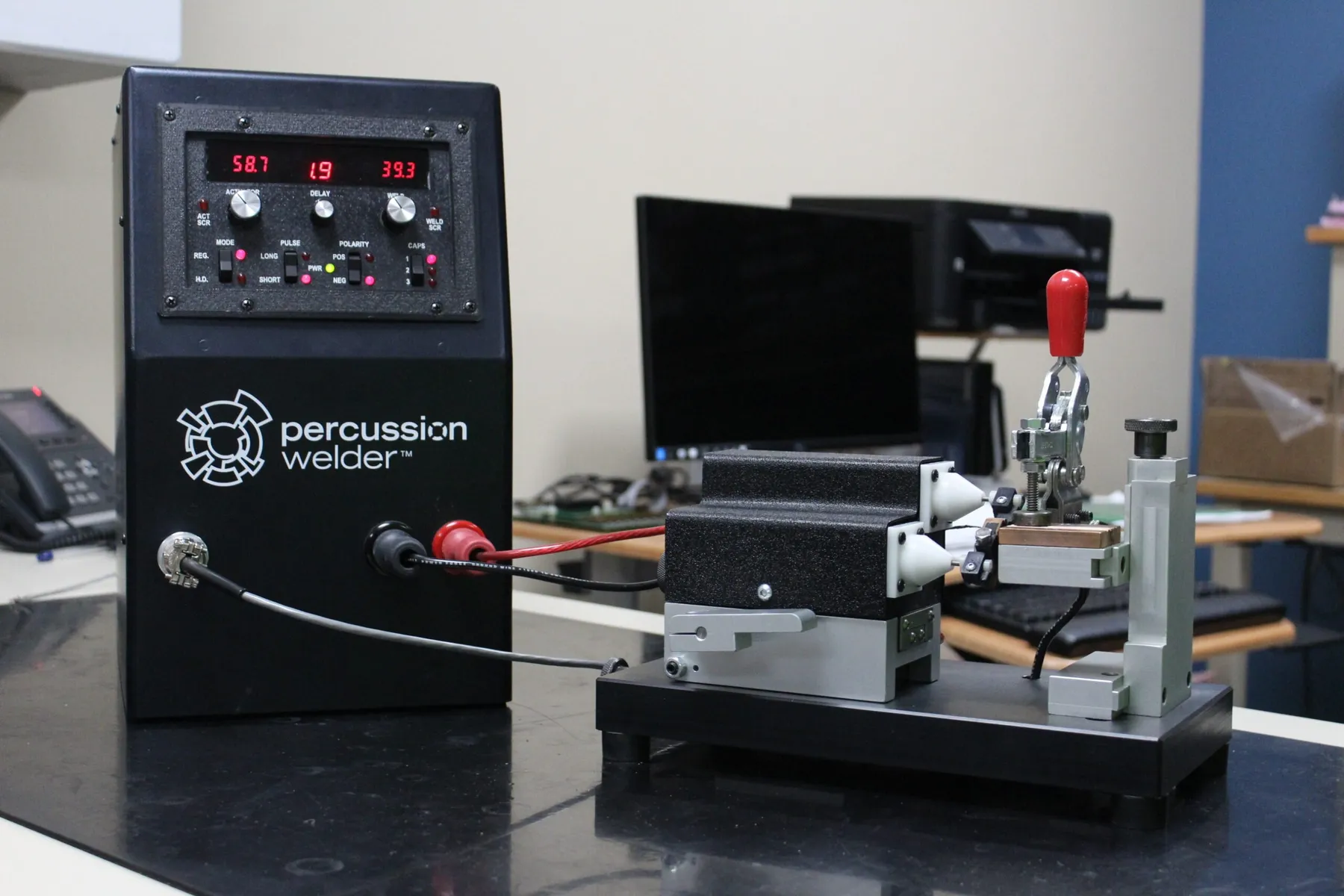 Percussion welder power supply with digital displays beside a bench fixture and laptop