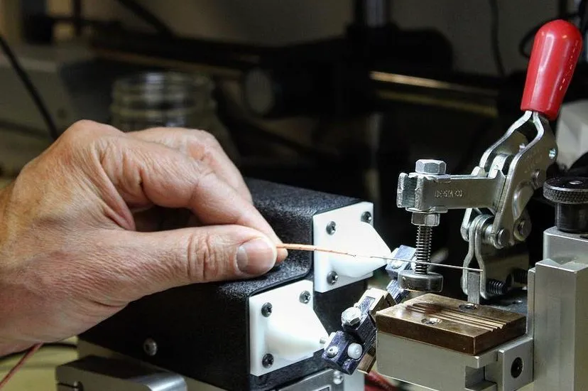 Hand placing a thin wire into the bench fixture next to the red toggle clamp